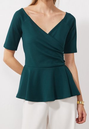 Femme portant un haut peplum vert foncé à manches courtes et col en V, associé à un pantalon blanc et un bracelet en or à son poignet droit.