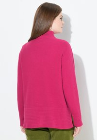 Donna con lunghi capelli castani, che indossa un maglione a collo alto rosa brillante e texturizzato e pantaloni verdi, di spalle contro un muro bianco.