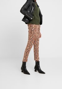 Person, der braune Schlangenleder-Hosen, schwarze Leder-Ankle-Boots, einen grünen Strickpullover und eine schwarze Lederjacke trägt, vor einem weißen Hintergrund.