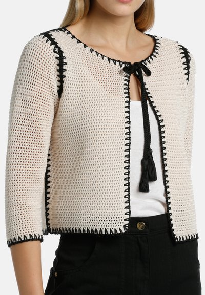 Neulecardigan vaaleassa sävyssä mustilla yksityiskohdilla. Ominaisuuksiin kuuluu pyöreä pääntie, kolmanneksen pituiset hihat, auki jätetty etuosa ja nauhakiristys tupsuilla.