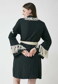 Svart klänning med långa klockärmar, prydd med vit frynsa vid ärmsluten och kragen. Har ett knytbälte och en avslappnad passform.