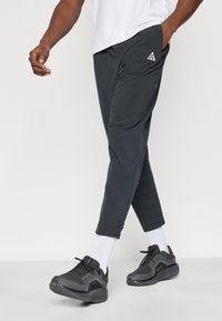 Στενόμενα γκρι σκούρα παντελόνια με διακριτική γυαλάδα, που διαθέτουν πλαϊνές τσέπες και λογότυπο. Συνδυασμένα με μαύρα αθλητικά παπούτσια και λευκές κάλτσες.