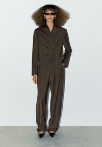 Brauner zweireihiger Blazer mit Schulterpolstern, kombiniert mit weit geschnittenen Hosen. Glattes Material und minimalistisches Design, ergänzt durch schwarze Sonnenbrillen.