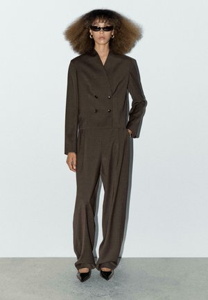 Bruine dubbelgeknoopte blazer met schoudervulling, gecombineerd met wijde broekspijpen. Gladde stof en minimaal ontwerp, met zwarte zonnebril.