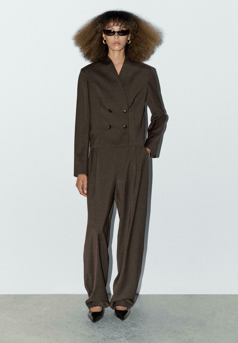 Brauner zweireihiger Blazer mit Schulterpolstern, kombiniert mit weit geschnittenen Hosen. Glattes Material und minimalistisches Design, ergänzt durch schwarze Sonnenbrillen.