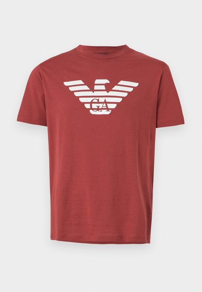 Camiseta de algodão vermelha com um logotipo de águia riscada horizontalmente em branco e "GA" ao centro. Gola redonda e mangas curtas.