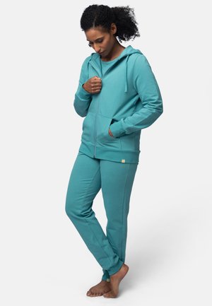 Felpa con zip turchese e pantaloni coordinati in un tessuto morbido. La felpa ha una tasca frontale e cordoncino, mentre i pantaloni presentano polsini alla caviglia.