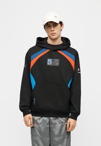 Jeune homme portant un sweat à capuche noir avec des accents bleus et orange, un pantalon gris et une casquette blanche Puma, debout devant un fond uni.