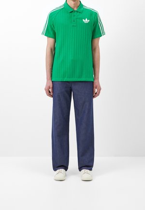 Polo vert côtelé avec trois bandes blanches sur les manches, arborant un petit logo blanc. Associé à un pantalon bleu ample et des chaussures blanches.