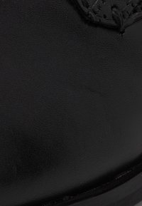 Chaussure en cuir noir présentant une texture lisse et un embout arrondi. Affiche des détails de couture complexes et une légère brillance à la surface.