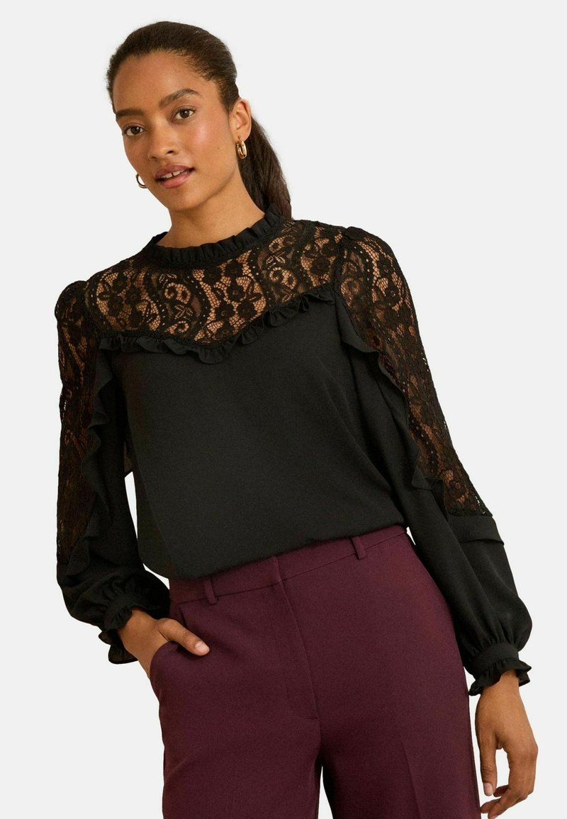 Fekete blúz csipke részletekkel, fodrokkal a vállakon és az ujjatlan mandzsettán, bordó nadrággal párosítva. Sima anyagfelület.