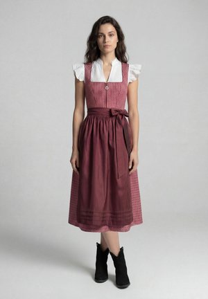 Jeune femme portant une robe traditionnelle dirndl bordeaux avec une blouse blanche en dentelle et des bottines noires, debout devant un fond gris uni.