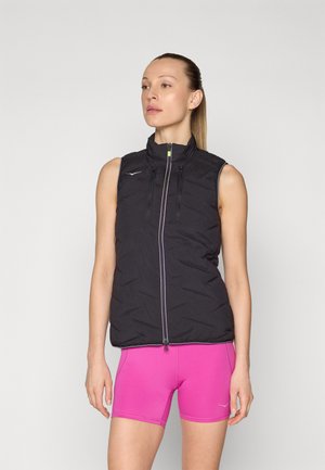 Svart ermeløs polstret vest med glidelåser og høy krage, parret med rosa shorts. Kjennetegn inkluderer et reflekterende logo og kontrasterende glidelåsdetaljer.