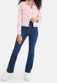 Femme portant une veste boutonnée rose clair, un haut blanc, un jean bleu foncé et des baskets blanches, debout avec une main près de l'ourlet de la veste.