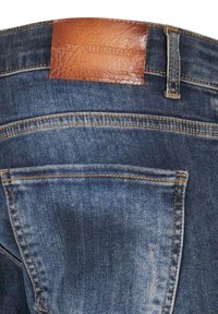 Jeans en denim bleu foncé avec une écusson en cuir marron texturé portant l'inscription "2Y PREMIUM", dotés de poches arrière standard et de coutures jaunes contrastées.