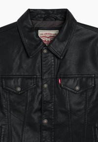 Veste en cuir noir au design classique, avec des boutons-pression, deux poches poitrine et une étiquette rouge sous la poche gauche. Matériau texturé.