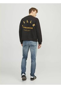 Sweatshirt noir avec du texte et des graphiques jaunes dans le dos, associé à un jean bleu et des baskets noires. Coupe et design décontractés.