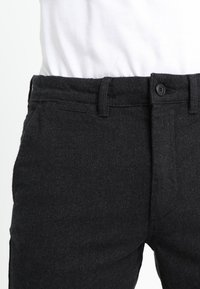 Mustat, teksturoidut housut kalanruotokuvioinnilla, joissa on nappisuljenta ja vyölenkit, osa asua, johon kuuluu valkoinen paita.