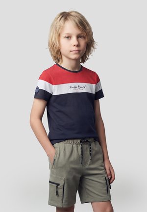 Niño rubio de pie con una mano en el bolsillo, vestido con una camiseta a rayas en rojo, blanco y azul marino y pantalones cortos cargo color oliva, sobre un fondo gris.