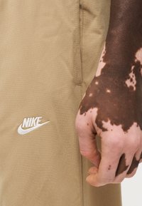 Μπεζ αθλητικά σορτς Nike με λευκό κεντητό λογότυπο. Το ύφασμα φαίνεται απαλό με ομαλή υφή και εμφανείς λεπτομέρειες ραφής.