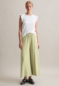 Weißes, geripptes, ärmelloses Oberteil mit Rüschenschultern und hellgrünen, weit geschnittenen Hosen. Schwarze Schuhe runden das Outfit ab.