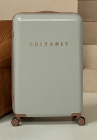 Harde koffer in lichtgrijs met gouden letters. Bevat ronde hoeken, een zijhandvat en vier wielen voor mobiliteit.