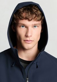 Marineblaue Kapuzenjacke mit glatter Textur, Frontreißverschluss und Kordelzugdetail. Kombiniert mit einem schwarz-weiß gestreiften Hemd.