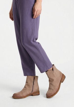 Person iført lilla bukser med lige ben og lysebrune ankelstøvler i ruskind med lynlås i siden, står på hvid baggrund.