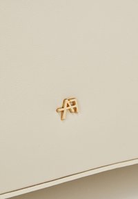 Krämfärgad läderyta med en guldig logotypaccent. Materialet har en slät textur med subtila kornmönster.