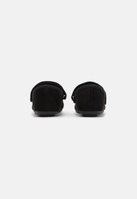 Pantofole in suède nero con profilo basso e cinturino regolabile a strappo, caratterizzate da una texture liscia e design a punta arrotondata.