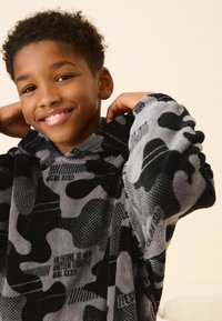 Pullover hoodie lavet af blødt fleece, med et sort og gråt camouflagemønster med varierede teksturer og trykte tekstdetaljer.