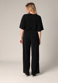 Jumpsuit noir avec des manches courtes et larges, tissu texturé avec un léger éclat, coupe ample, taille élastique et design de jambe droite.