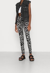 Gilet senza maniche in denim nero, crop top bianco, leggins con stampa a righe zebrate e sandali neri. I leggins presentano righe alternate in bianco e nero.