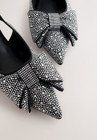 Čierne špicaté topánky zdobené trblietavými kryštálikmi a veľkými dekoračnými mašľami na svetlom pozadí.