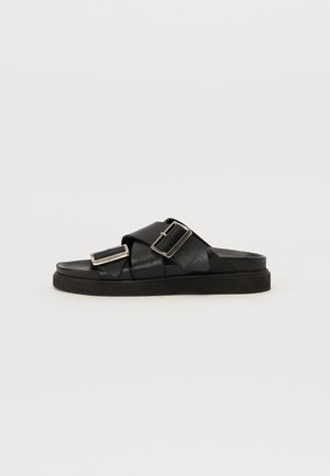Sandales en cuir noir avec un large design de bride en croix, comportant deux boucles métalliques et une semelle en caoutchouc plate pour le soutien.