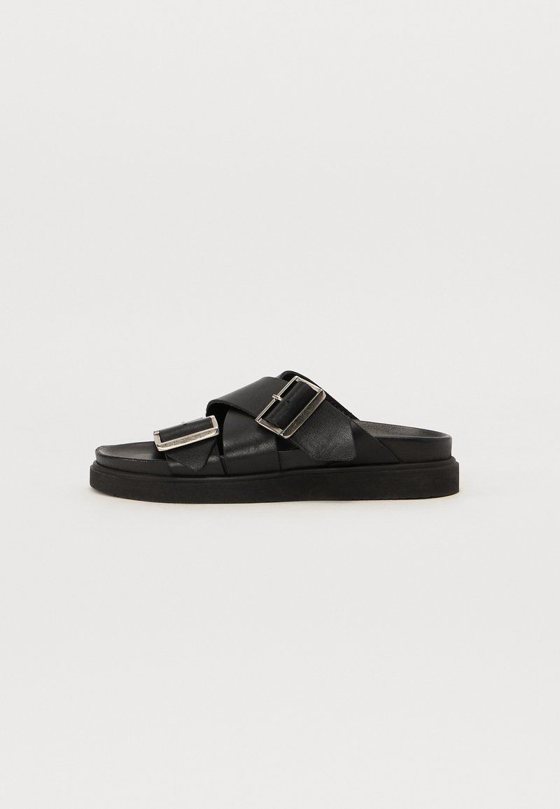 Black leather sandals with a wide cross strap design, featuring two metallic buckles and a flat rubber sole for support.