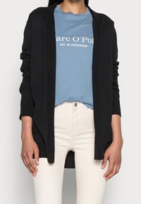 Cardigan nero con design aperto, maniche lunghe e texture morbida, indossato sopra una maglietta azzurra chiara e pantaloni bianchi.