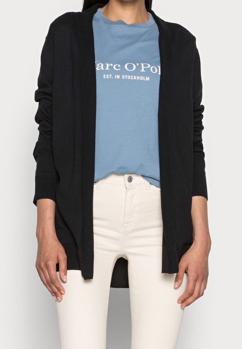 Cardigan nero con design aperto, maniche lunghe e texture morbida, indossato sopra una maglietta azzurra chiara e pantaloni bianchi.