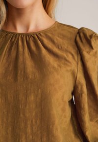 Ockergelbe Bluse mit strukturiertem Oberflächen, rundem Halsausschnitt und gerafften Details an den Schultern, mit langen Ärmeln und subtiler Volumen.