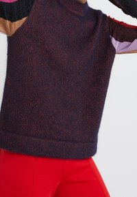 Maglione lavorato a maglia in un blu navy scuro con una finitura testurizzata, caratterizzato da maniche multicolori con filo metallico in rosa, rosso e nero.