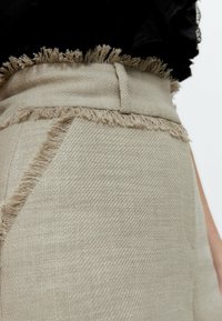 Béžové kalhoty s roztrženým lemem, vyrobené z texturované látky. Mají úzký pas a jemný rybí vzor.