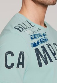 Jasnoniebieska bawełniana koszulka z dużym ciemnogranatowym napisem i grafikami na ramieniu oraz klatce piersiowej, z okrągłym dekoltem i krótkimi rękawami.