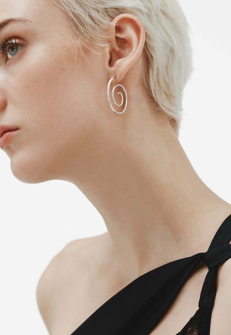 Aro de plata elegante con un diseño en espiral, llevado en la oreja de una modelo. Suave y corto cabello rubio contrasta con una blusa negra.