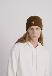 Bruine geribde gebreide muts met oranje logodetail, gedragen door een persoon in een witte hoodiekleding met reliëf merknaam.