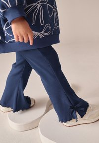Pantalon côtelé bleu marine avec ourlets évasés et détails à volants, associé à des baskets de couleur claire. La main du mannequin pointe vers le design.