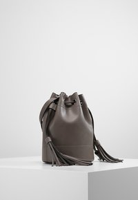 Harmaasta nahasta valmistettu bucket-laukku, pyöreä muoto, nyörikiinnitys ja pitkät hapsut. Sileä pinta, jossa hienovaraiset ompeluaksentit.