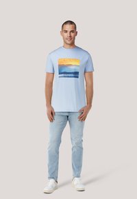 Camiseta de algodón azul claro con un gráfico rectangular que muestra montañas y agua, etiquetada "GANT USA Sailing". Llevada con jeans claros y zapatillas blancas.