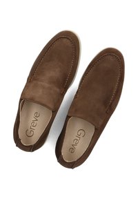 Mocassins en daim marron avec un design à enfiler, présentant des détails de couture et une semelle en caoutchouc de couleur claire. L'intérieur affiche le logo "Greve".