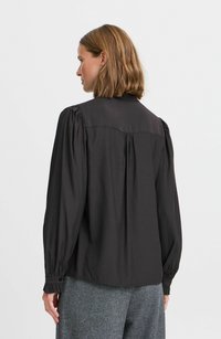 Schwarze Bluse mit langen Ärmeln, gefältelten Schultern und geknöpften Manschetten. Subtile Glanz, gerader Schnitt und ein kleiner Kragen. Der Stoff ist glatt.