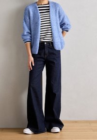 Un cardigan en tricot bleu clair porté sur une chemise à rayures noires et blanches, associé à un jean large bleu foncé et des baskets blanches.